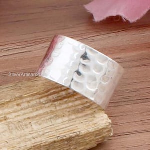 May include: A wide, hammered silver ring with a textured surface. The ring is set against a wooden background, with a pink flower petal in the upper right corner. The ring's surface reflects light, highlighting its hammered design.