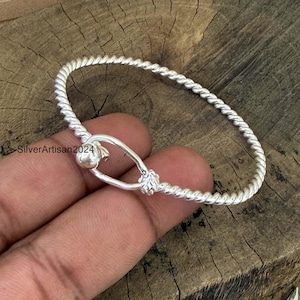 Puede incluir: Una pulsera de plata con un diseño de cuerda trenzada y un mecanismo de cierre central en forma ovalada. La superficie metálica es pulida y reflectante. El texto SilverArtisan2024 es visible en el lado izquierdo cerca del cierre.