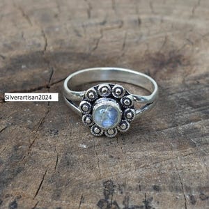 May include: A silver ring featuring a central round moonstone cabochon surrounded by a scalloped floral-shaped metal border with small raised dots. The band is split at the shoulders. The text Silverartisan2024 is visible on the left side.