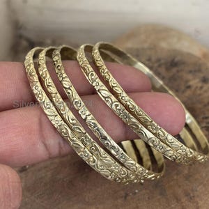 Puede incluir: Un conjunto de cuatro brazaletes de metal en tono dorado con un patrón continuo de flores y enredaderas en relieve. Cada pulsera circular tiene un acabado pulido con intrincados detalles tallados que recorren toda la circunferencia.