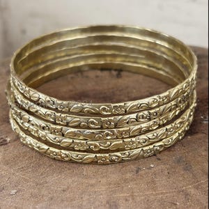 Może przedstawiać: Zestaw czterech złotych metalowych bransoletek typu bangle z ciągłym tłoczonym wzorem kwiatów i pnączy. Każda okrągła bransoletka ma polerowane wykończenie ze szczegółowymi rzeźbieniami przedstawiającymi małe kwiaty i liście.