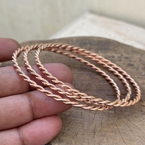Può includere: Tre braccialetti color rame con un design a corda intrecciata. I braccialetti sono impilati, mostrando la loro trama intricata e la calda tonalità metallica. I braccialetti sono esposti su uno sfondo di legno naturale.