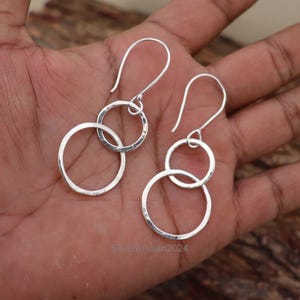 Könnte beinhalten: Ein Paar silberne Ohrringe mit Haken-Design. Jeder Ohrring hat drei miteinander verbundene Kreise unterschiedlicher Größe. Die Ohrringe werden vor einem neutralen Hintergrund präsentiert, der ihren metallischen Glanz und ihr schlichtes, elegantes Design hervorhebt.