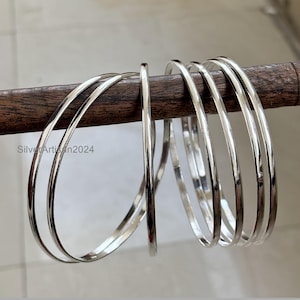 Op de afbeelding: Een verzameling glanzende, zilveren armbanden. De dunne, ronde armbanden worden getoond op een donkere houten staaf. De afbeelding benadrukt het reflecterende oppervlak van de sieraden. De tekst "Silver Artisan2024" is zichtbaar in de linker benedenhoek.