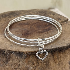 May include: Three silver bangles with a hammered texture and a heart charm. The bangles are stacked on a wooden surface. The heart charm is a simple open design.