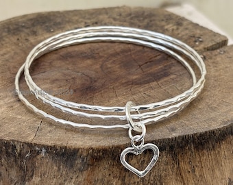 Pode incluir: Três pulseiras de prata com textura martelada e um berloque de coração. As pulseiras estão empilhadas sobre uma superfície de madeira. O berloque de coração tem um design simples e aberto.