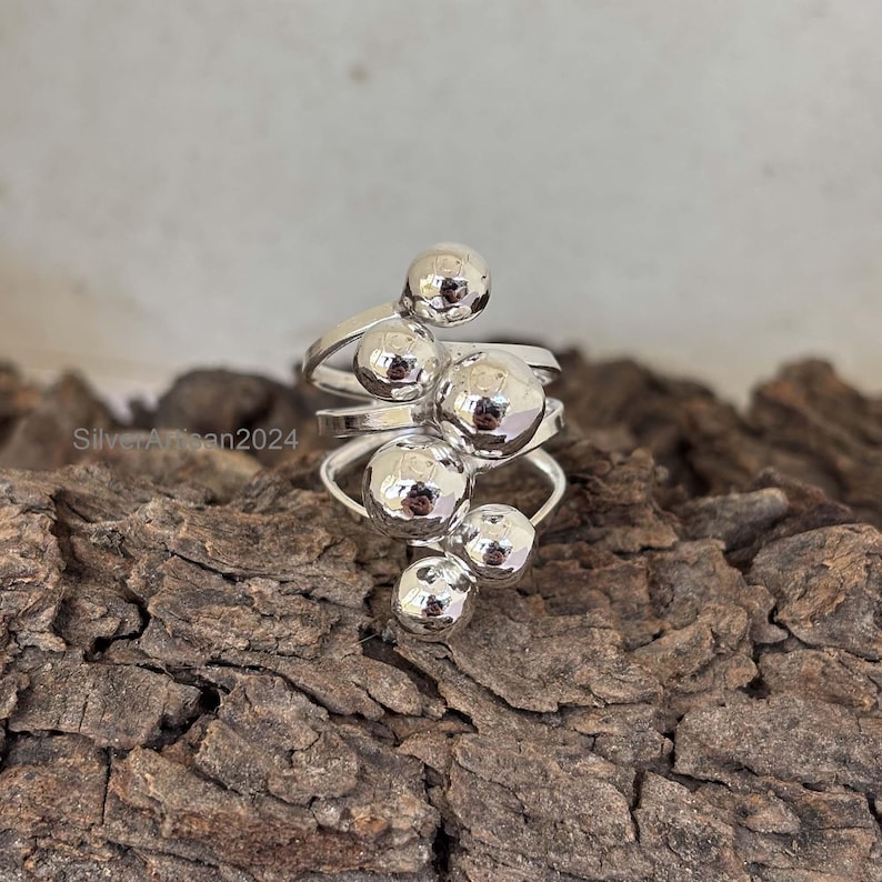 May include: A silver ring with a cluster of five round, textured spheres. The spheres are arranged in a cascading design, with the largest sphere at the top and the smallest sphere at the bottom.