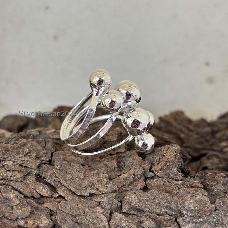 May include: A silver ring with multiple bands and round silver beads. The ring is on a wooden surface.