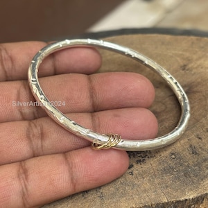 May include: A silver bangle bracelet with a textured surface and a cluster of three gold-colored rings. The bracelet is held in a hand and is displayed on a wooden surface. The text "SilverArtisan2024" is visible.