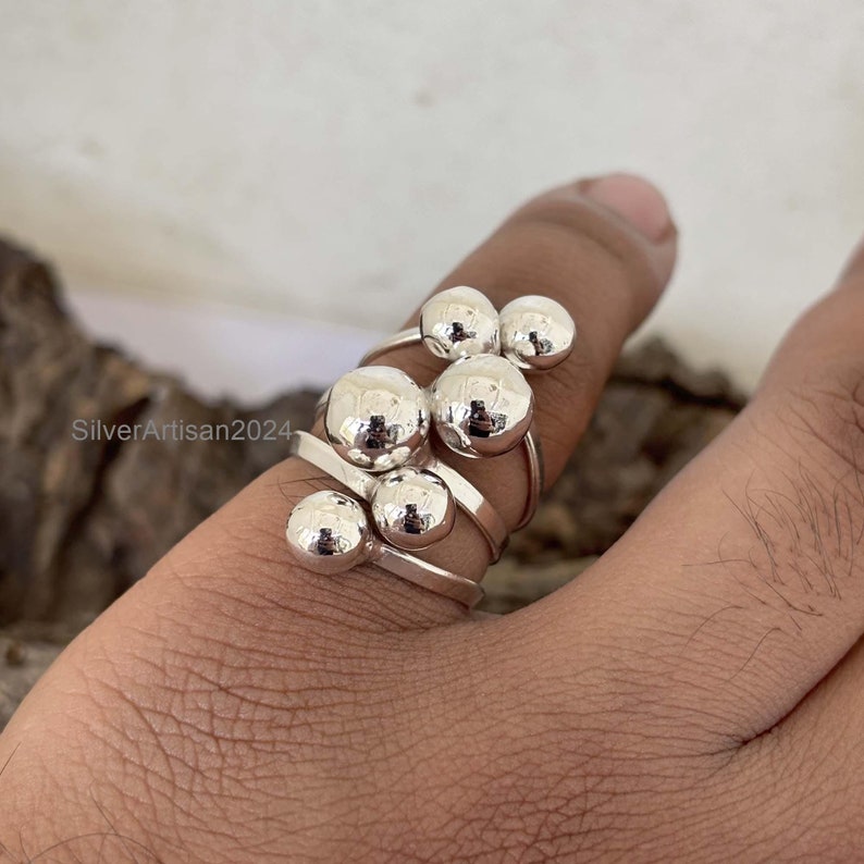 May include: A silver ring with multiple round balls attached to a thin band. The ring is worn on a finger.