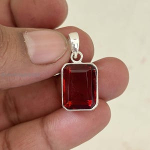 May include: A rectangular, deep red gemstone pendant with a silver-colored setting. The pendant is held by a hand, showcasing the polished, faceted surface of the gem. The pendant's shape is an elongated rectangle.
