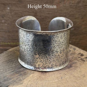 Op de afbeelding: Een zilveren manchetarmband met een reliëf bloemmotief. De armband is 5 cm hoog. De manchet wordt gepresenteerd op een houten oppervlak.
