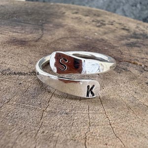 May include: A silver-colored bypass ring featuring two flat, rounded ends. The top end is stamped with the letter S, and the bottom end is stamped with the letter K. The metal surface has a polished, reflective finish with visible text SilverArtisan2024.