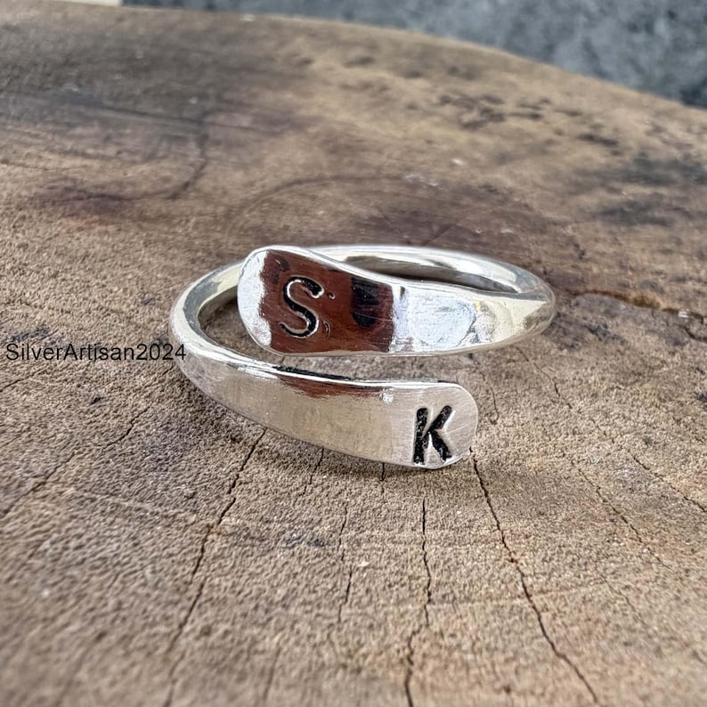 May include: A silver-colored bypass ring featuring two flat, rounded ends. The top end is stamped with the letter S, and the bottom end is stamped with the letter K. The metal surface has a polished, reflective finish with visible text SilverArtisan2024.