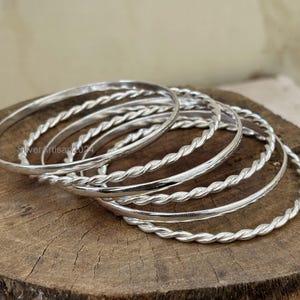 May include: A collection of silver bangles with a twisted rope design. The bangles are displayed on a wooden surface. The silver bangles have a shiny, reflective surface. The image is well-lit, highlighting the details of the jewelry.