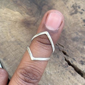 May include: A silver ring with a V-shaped design. The ring is worn on a finger, showcasing its minimalist and modern style. The ring's design is simple yet elegant, suitable for everyday wear. The ring is made of a shiny, metallic material.