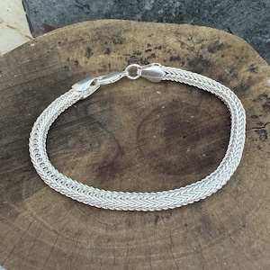 Può includere: Un bracciale in argento caratterizzato da un design a catena intrecciata e tessuta con finitura lucida. Il bracciale è fissato con un meccanismo a moschettone, che mostra un motivo a maglie coerente e complesso lungo tutta la lunghezza del gioiello.