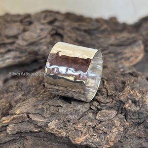 Op de afbeelding: Een brede, gehamerde zilveren ring. De ring heeft een getextureerd oppervlak en een gepolijste afwerking. De ring wordt getoond op een stuk hout. De tekst "SilverArtisan2024" is zichtbaar op het hout.