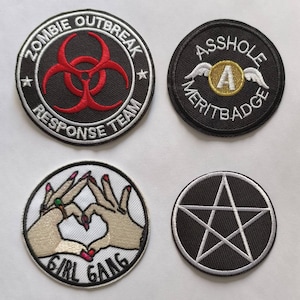 May include: Four embroidered patches with different designs. One patch says "ZOMBIE OUTBREAK RESPONSE TEAM" with a red biohazard symbol. Another says "ASSHOLE MERIT BADGE" with a winged "A". A third patch shows hands forming a heart with "GIRL GANG". The last patch has a white pentagram.