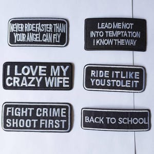 May include: A collection of six black and white embroidered patches with various phrases. The patches include text such as "Never ride faster than your angel can fly," "I love my crazy wife," and "Fight crime shoot first."