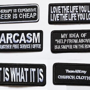 Op de afbeelding: Zes zwarte en witte geborduurde patches met verschillende spreuken. De patches omvatten "Therapie is duur, bier is goedkoop", "Sarcasme, nog een gratis service", "Leef het leven dat je leeft, leef het leven dat je liefhebt", "Mijn idee van "hulp van boven" is een scherpschutter op het dak", "Het is wat het is", en "Dit zijn mijn kerk-kleding".