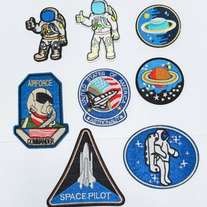 May include: Nine embroidered iron-on patches featuring space and astronaut themes. The patches are blue, red, white, black, and grey. The patches include images of astronauts, spaceships, planets, and stars. The patches have text that reads "Airforce Commander", "United States of America Astronaut", "Space Pilot", and "Airforce".