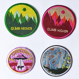 Pode incluir: Quatro patches bordados para passar a ferro. Dois patches mostram uma cordilheira com um sol e o texto "CLIMB HIGHER". Um patch mostra um OVNI rosa com um raio de luz que brilha sobre uma vaca. O último patch mostra um design abstrato azul e laranja.