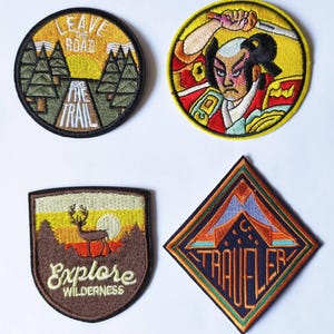May include: Four embroidered patches with different designs. The first patch is green and yellow with the text "Leave the Road Take the Trail". The second patch is yellow and red with a samurai warrior design. The third patch is brown with a deer silhouette and the text "Explore Wilderness". The fourth patch is a diamond shape with a mountain design and the text "Traveler".
