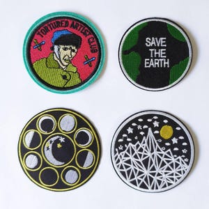 Op de afbeelding: Vier geborduurde strijkpatches. De eerste patch is groen, rood en blauw met een cartoon van een man met een blauwe hoed en de tekst "Tortured Artist Club". De tweede patch is zwart met groene tekst die "Save the Earth" zegt. De derde patch is zwart met een gouden rand en een witte halve maan met sterren. De vierde patch is zwart met een witte rand en een wit berggebied met een gele zon en sterren.