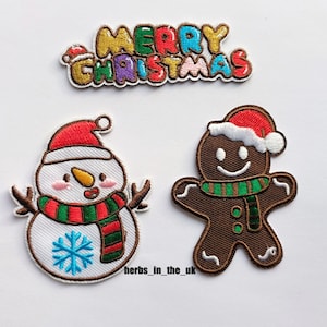 Pode incluir: Três patches bordados para passar a ferro que representam um boneco de neve, um homem de gengibre e a frase "Feliz Natal". O boneco de neve é branco com um cachecol vermelho e verde e um floco de neve azul. O homem de gengibre é castanho com um chapéu vermelho e branco e um cachecol verde. A frase "Feliz Natal" está em letras coloridas.
