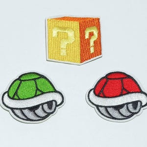 May include: Three embroidered patches featuring a green and silver, a red and silver, and a yellow and orange question mark block. The patches are all from the Super Mario Bros. video game series.