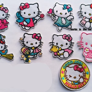 May include: A selection of embroidered Hello Kitty patches. The patches depict Hello Kitty in diverse outfits, such as a princess dress, fairy wings, and a witch's hat. One patch is circular, featuring the text "GOOD JOB!" and "HELLO KITTY".