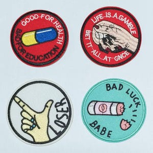 May include: Four embroidered iron-on patches with different designs and sayings. The first patch is red with white and blue pill and the text "GOOD FOR HEALTH BAD FOR EDUCATION". The second patch is red with a hand holding a small white object and the text "LIFE IS A GAMBLE BET IT ALL AT ONCE". The third patch is white with a hand pointing a finger and the text "LOSER". The fourth patch is turquoise with a broken pink pill and the text "BAD LUCK BABE".
