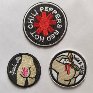 Puede incluir: Tres parches bordados. El primer parche es negro con texto blanco que dice "Red Hot Chili Peppers" y una explosión de estrella roja. El segundo parche es negro con texto blanco que dice "Spank Me" y una huella de mano rosa sobre un fondo beige. El tercer parche es negro con texto blanco que dice "Luca Taco" y una ilustración roja y amarilla de una persona recibiendo un taco.