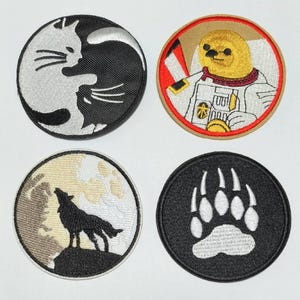May include: Four embroidered iron-on patches. The first patch is black and white with a yin yang design featuring a cat. The second patch is beige with a red border and features a cartoon astronaut wearing a spacesuit. The third patch is beige and black with a wolf howling at the moon. The fourth patch is black with a white bear paw print.