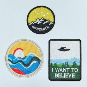 Può includere: Tre toppe ricamate da stirare. La prima toppa è gialla con montagne nere e il testo "WANDERER". La seconda toppa è bianca con una scena oceanica blu e gialla. La terza toppa è bianca con un bordo verde e il testo "I WANT TO BELIEVE" e un UFO nero.