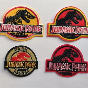 May include: Four embroidered iron-on patches featuring the Jurassic Park logo. The patches are yellow, red, and beige with black and white embroidery. The logos feature a dinosaur skeleton and the text "Jurassic Park".