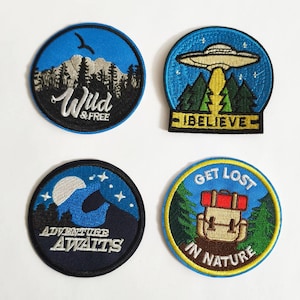 Op de afbeelding: Vier geborduurde patches met natuurthema's. De eerste patch is blauw met witte tekst die "Wild & Free" zegt en een bergketen met bomen afbeeldt. De tweede patch is blauw met witte tekst die "I Believe" zegt en een vliegende schotel boven een bos afbeeldt. De derde patch is zwart met witte tekst die "Adventure Awaits" zegt en een maan en sterren boven een bergketen afbeeldt. De vierde patch is blauw met gele tekst die "Get Lost in Nature" zegt en een rugzak in een bos afbeeldt.