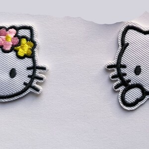 May include: Two white embroidered iron-on patches of Hello Kitty. One patch features Hello Kitty with a pink and yellow flower on her head. The other patch features Hello Kitty with a pink flower on her head and a pink bow on her arm.