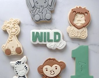 May include: A set of nine decorated cookies featuring a giraffe, elephant, lion, monkey, zebra, and a number one. The cookies are decorated with green, brown, and white icing. The cookies are arranged on a white marble surface. The cookies say "WILD one" on one of the cookies.