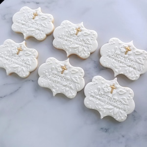 May include: Six white cookies decorated with gold icing and the words "Jasmines First Holy Communion".