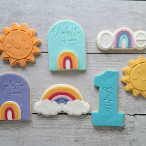 May include: A set of six decorated sugar cookies featuring a rainbow theme. The cookies are decorated with rainbows, clouds, suns, and the words "Violetta is one".