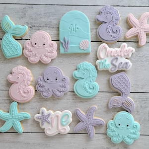 May include: A collection of decorated sugar biscuits with a sea theme. The biscuits feature mermaid tails, octopuses, seahorses, and starfish in pastel colours. Some biscuits have text that reads "Isla" and "One der the Sea".