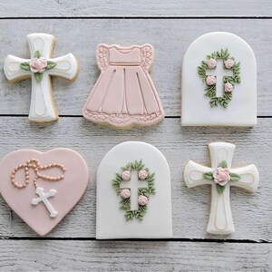 May include: A selection of decorated sugar biscuits in the shape of a cross, dress, heart, and arched plaque. The biscuits are white and pink, with floral and cross designs. They are arranged on a light wood surface.