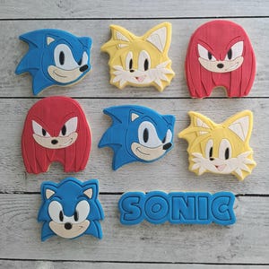 May include: A selection of character-shaped biscuits featuring Sonic the Hedgehog and friends. The biscuits are blue, yellow, and red, with white and tan details. The word "SONIC" is spelled out in blue biscuit letters.