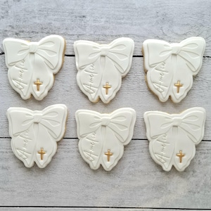 May include: Six white bow-shaped biscuits with gold crosses and the name "Amelia" written on them. The biscuits are arranged on a light wood-grain surface. The biscuits have a detailed bow design.