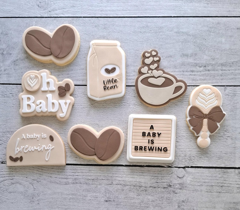 May include: Assortment of baby shower cookies in the shape of coffee beans, a milk carton, a coffee cup with hearts, a rattle, and signs that say "Oh Baby" and "A baby is brewing". The cookies are decorated in shades of brown and beige.
