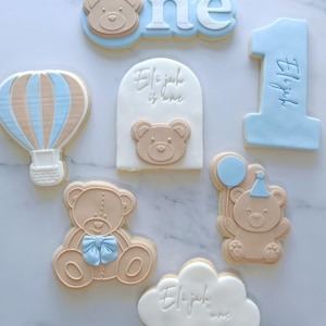 May include: A set of nine decorated sugar cookies for a first birthday. The cookies feature a blue and brown hot air balloon, a blue number one, a cloud with the text "Elijah is one", a teddy bear with a blue bow, a teddy bear with a blue balloon and party hat, and two teddy bear cookies.
