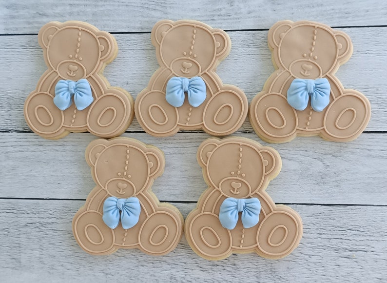 May include: Five brown teddy bear-shaped sugar cookies decorated with light blue bows. The cookies are arranged in a row on a light brown wooden surface.