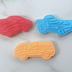 Pu&ograve; includere: Tre biscotti di zucchero decorati a forma di auto da corsa. I biscotti sono rossi, blu e arancioni.